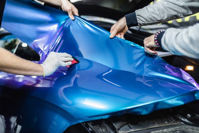 Two installers stretching blue chrome vinyl over a hood